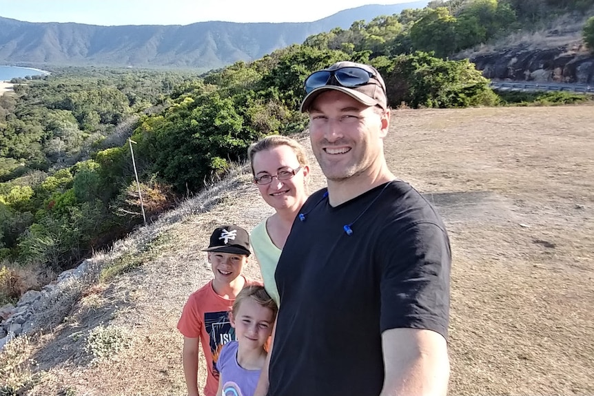 A group selife photo of Josh, his wife, and two young children, on top of a hill overlooking bushland and a beach.
