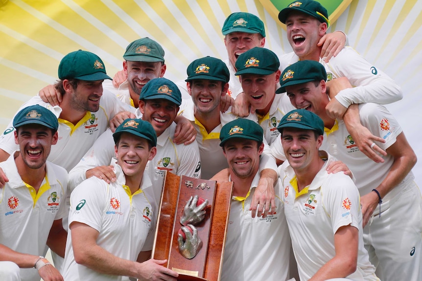 The Australian players crowd around the trophy while smiling and celebrating.
