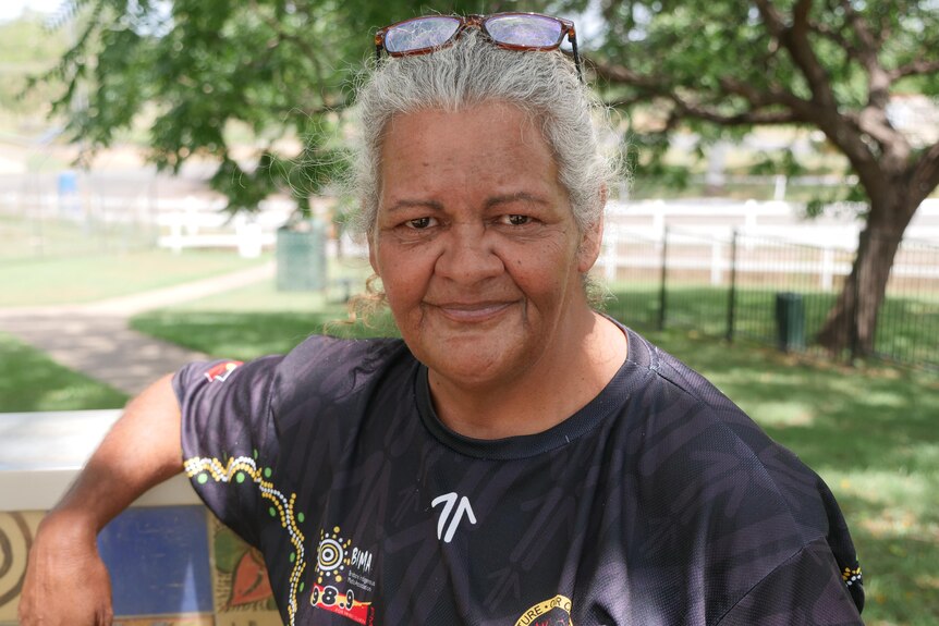 A smiling Indigenous woman with grey hair leans against a sign in a park.