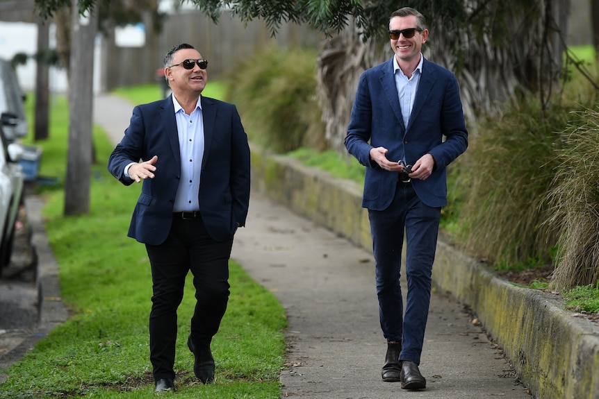 two men wearing dark sunglasses walking outside on a footpath and smiling