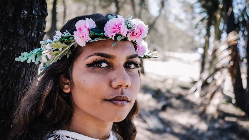 A photo of an Indigenous teenager wearing a floral crown looking at the camera standing outdoors.