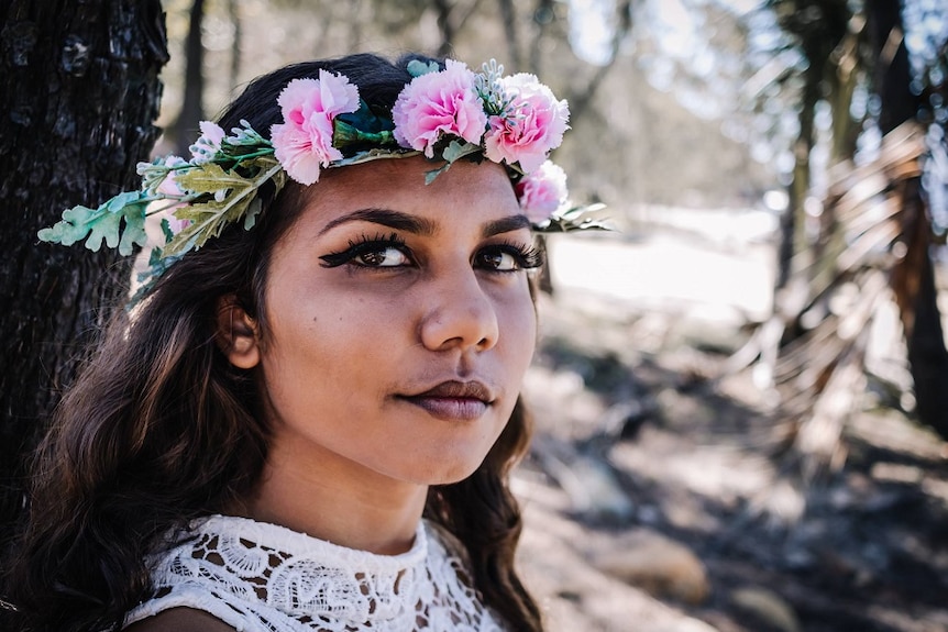 A photo of an Indigenous teenager wearing a floral crown looking at the camera standing outdoors.