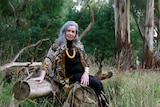 A woman with long silver hair wearing big yellow beads sits on a log in a beautiful clearing