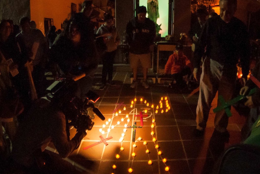 A group gathers at night around an outline of a body on the ground, illuminated by candles for the missing 43 Mexican students.