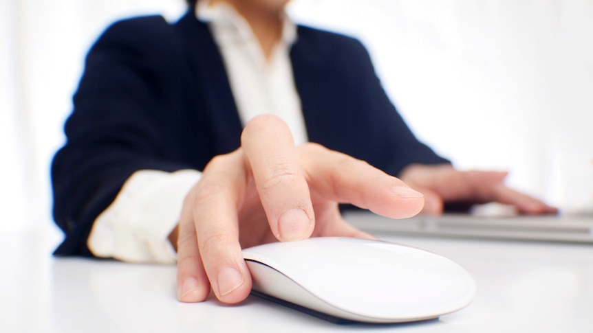 A faceless person wearing a shirt and jacket clicks on a mouse on a desktop.