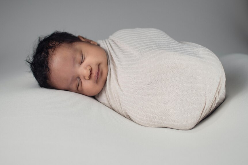 A black-haired baby wrapped in a white blanket sleeping on a bed with white linen. 