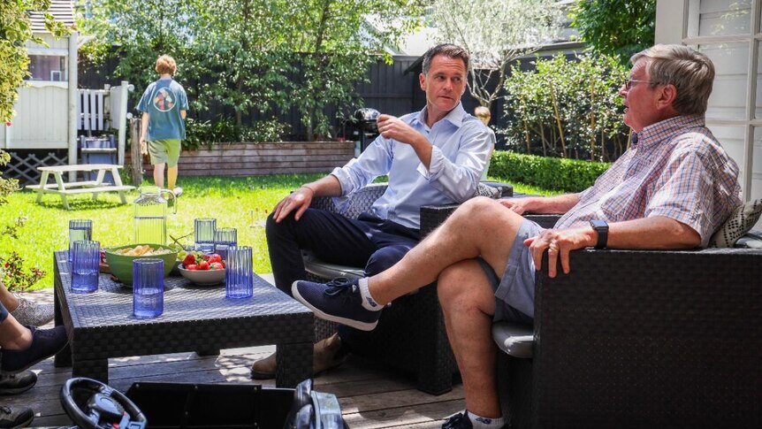 An older man sits with an older, but younger than him, man in an open-layout outdoor lounge