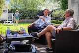 An older man sits with an older, but younger than him, man in an open-layout outdoor lounge