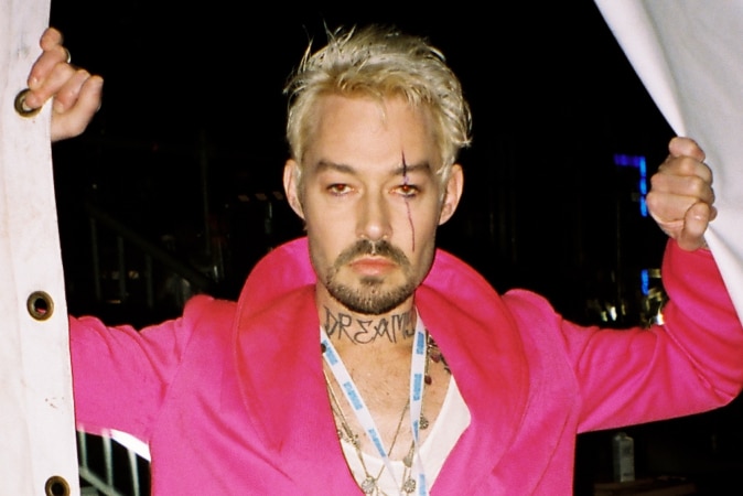 Daniel Johns stands holding open two sides of a large tent wearing a bright pink coat with bleached hair