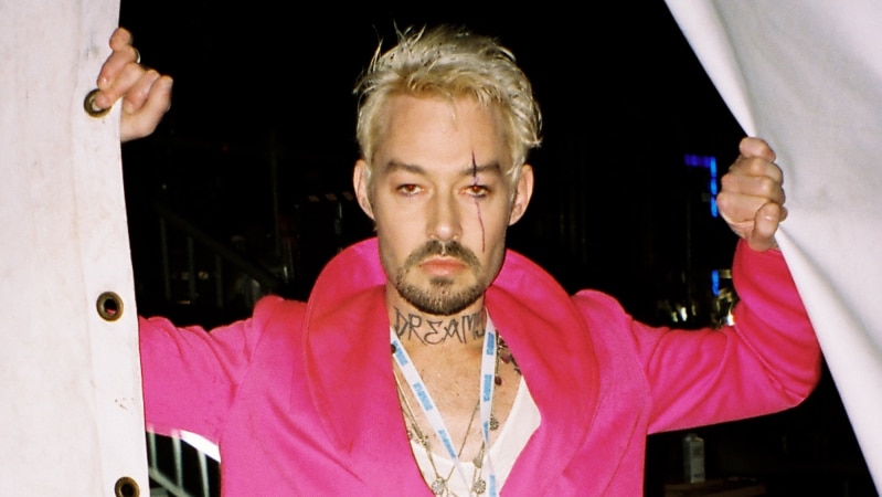 Daniel Johns stands holding open two sides of a large tent wearing a bright pink coat with bleached hair