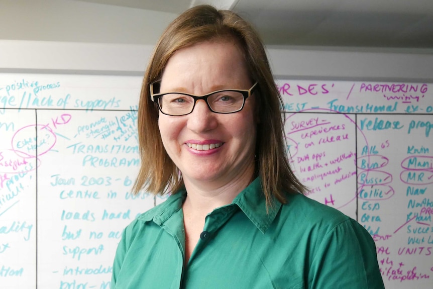A woman with short brown hair, glasses and a green dress stands in front of a whiteboard.