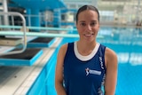 Diver Anabelle Smith, wearing a blue singlet, is standing on pool deck with diving boards behind her, smiling at the camera. 
