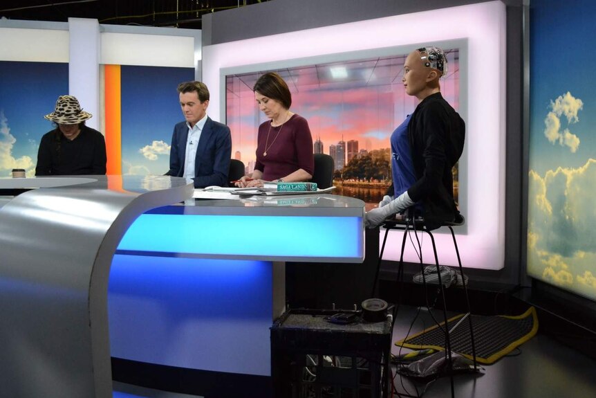 The torso of Sophia the robot sits on a stool at the news desk alongside the presenters and her creator.