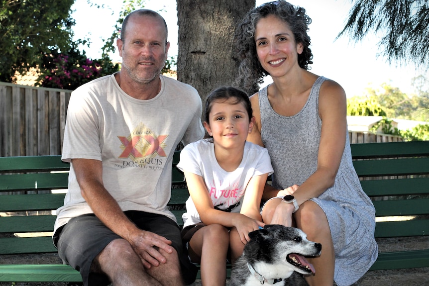A family of three sit on a park bench with their dog