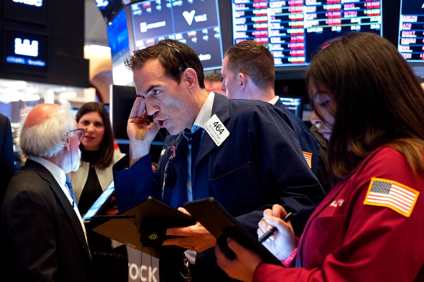 A man yells into a telephone headpiece while standing in front of a screen showing market information