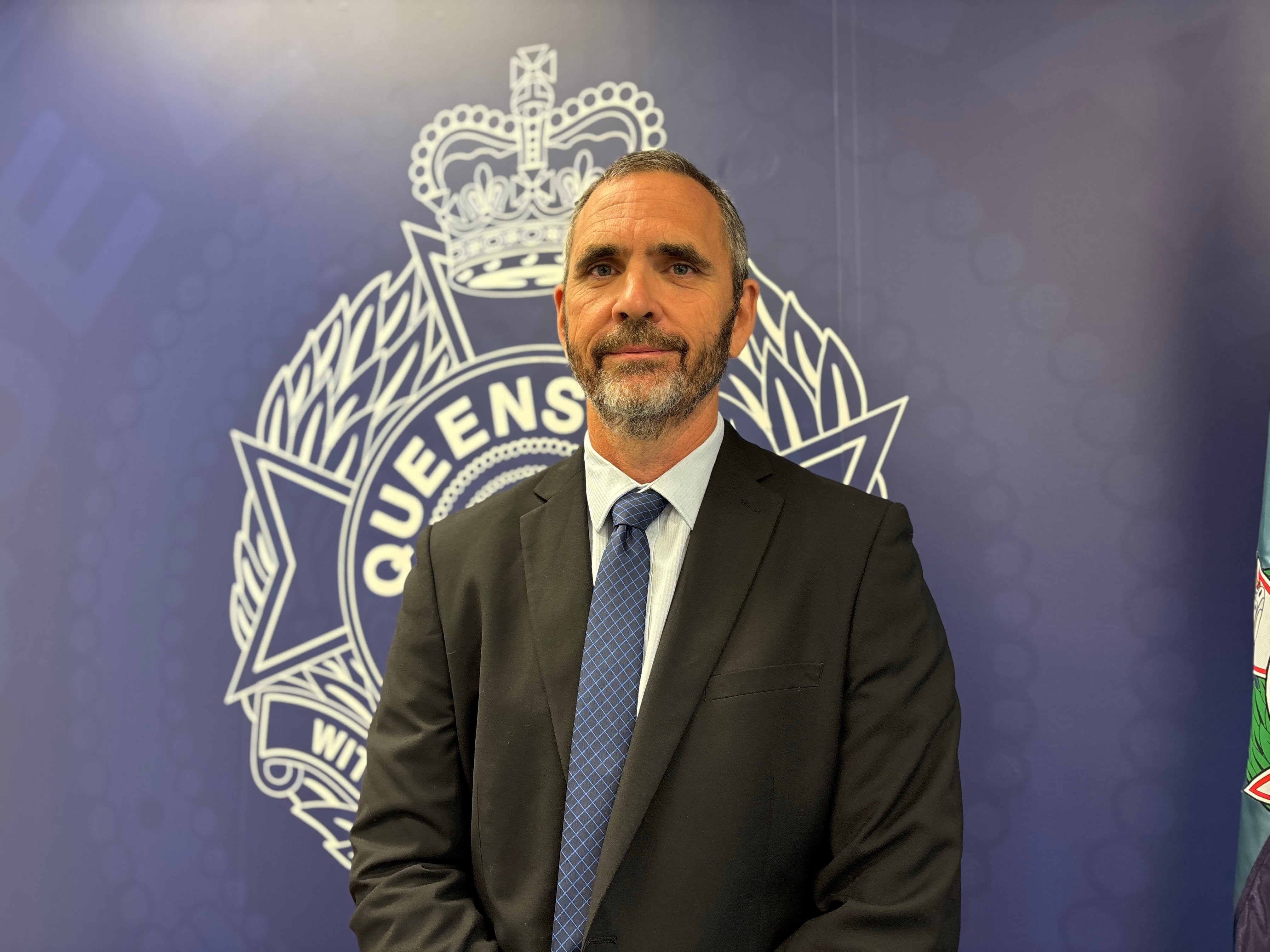A plain-clothes detective in a suit standing in front of a Queensland Police emblem.