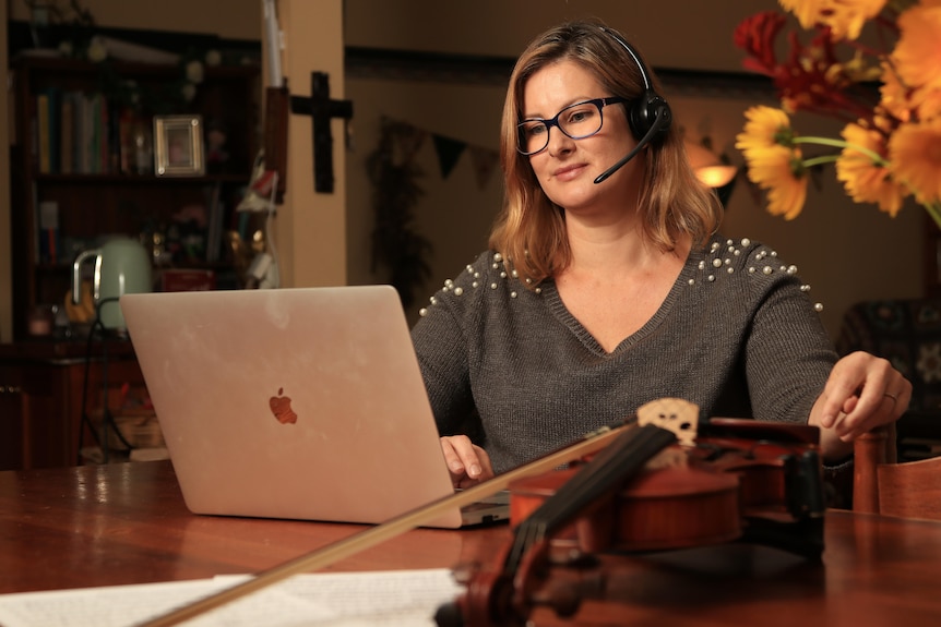 Sarah Moir sits at her kitchen table wearing a phone headset with a violin on the table and laptop.