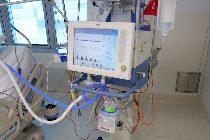 A ventilator with a screen and lots of tubes and cords connected to it sits beside an empty hospital bed.