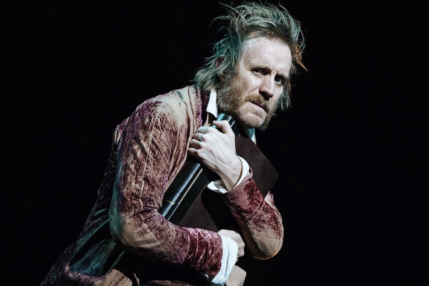 On stage, a blond, scruffy-haired, middle-aged man in Victorian era dress looks disgruntled, holding a large book to his chest