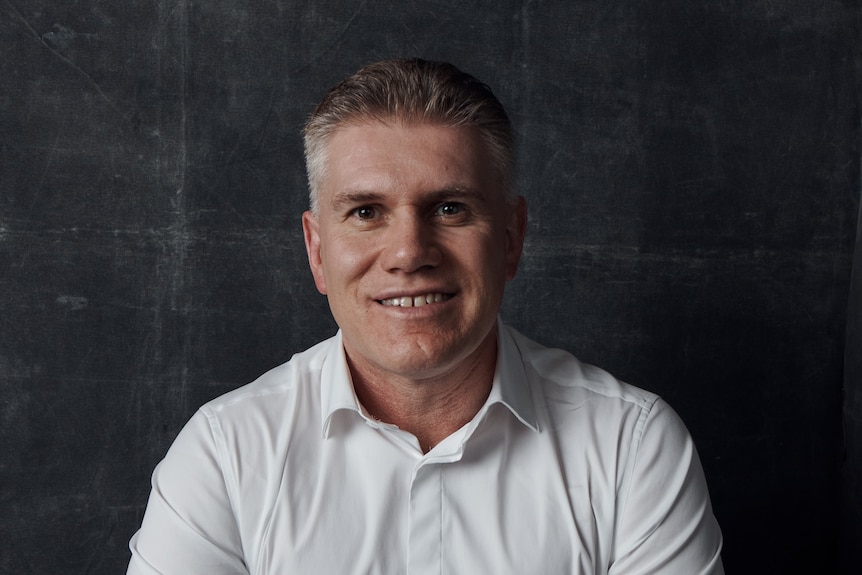 A middle-aged man smiles at the camera. He has a white short, a gap tooth smile, brown eyes and greying brown hair.