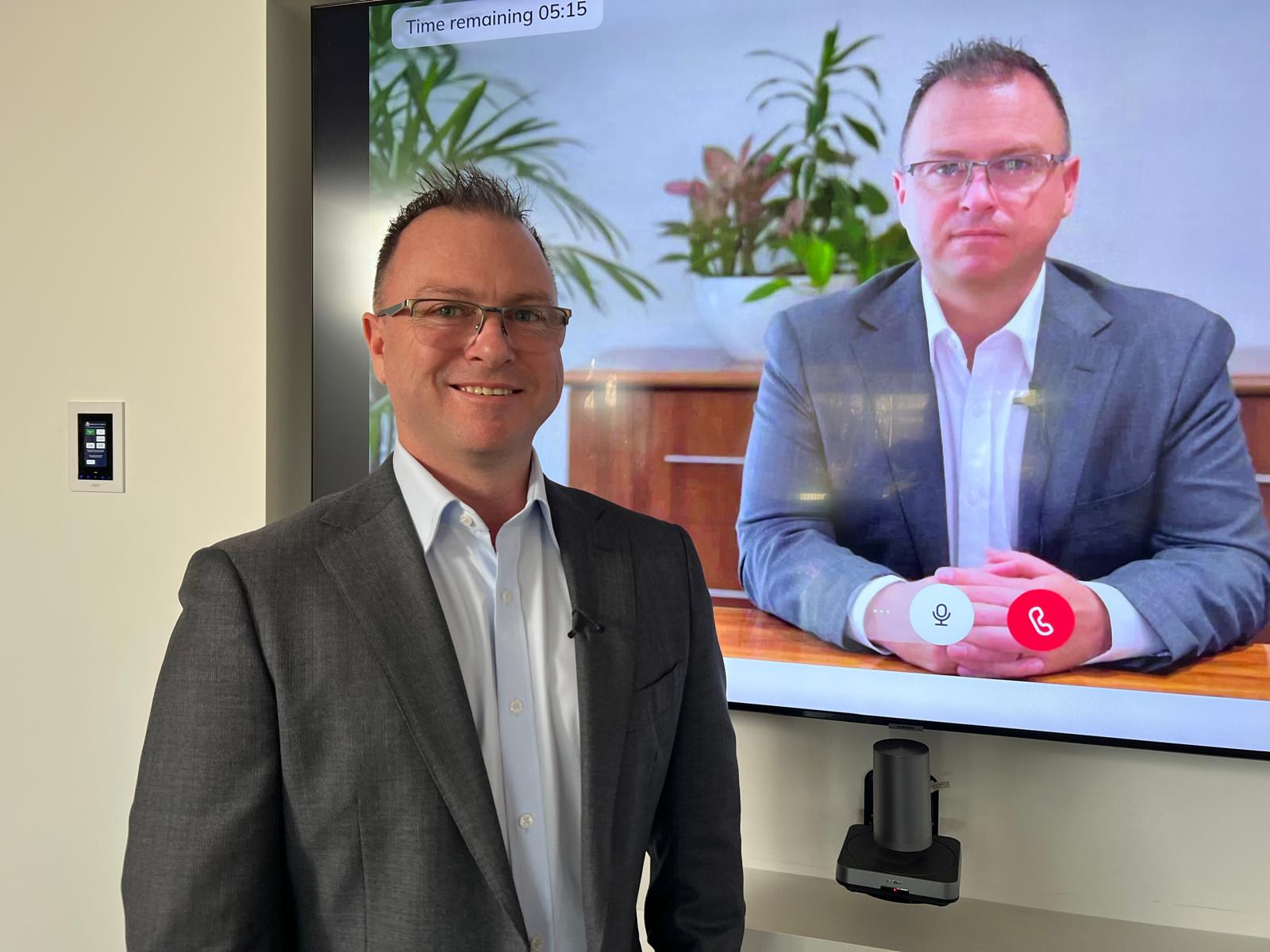 A man stands in front of a TV screen projecting a deepfake version of himself at a desk.