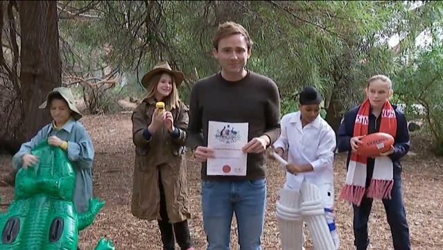Presenter holds citizenship certificate, children behind dressed in typical Australian garb
