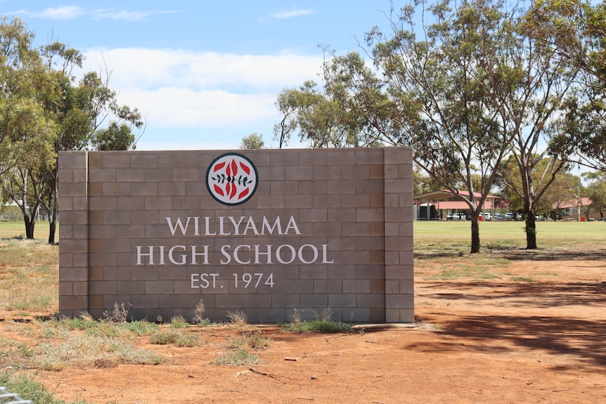 A stone sign that says Willyama high school est. 1974 on some dirt ground and in front of some trees, a grass patch and building