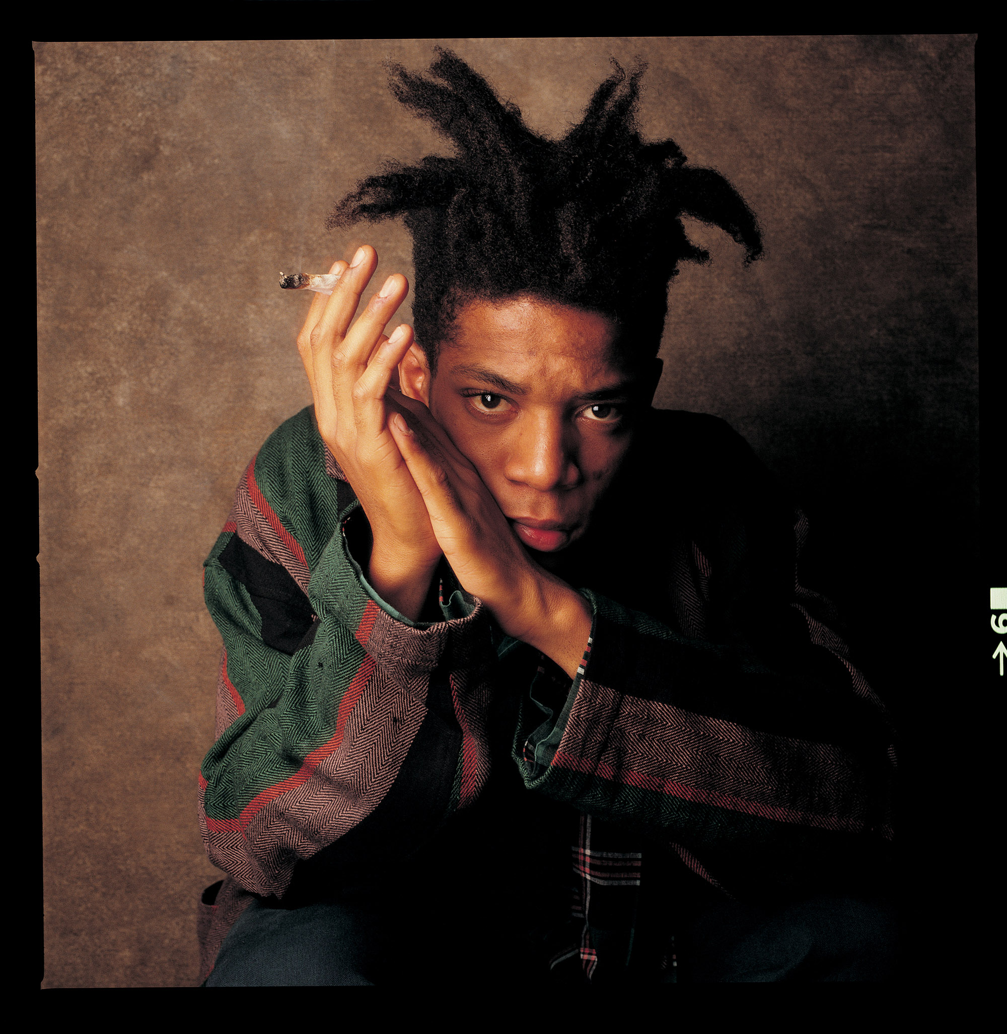 Square colour photograph of artist Jean-Michel-Basquiat leaning forward into hands clasped in front of photograph backdrop.