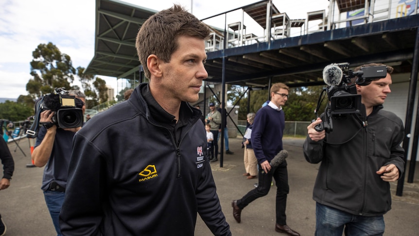 An Australian cricketer walks out of a cricket ground surrounded by TV cameramen after his game is called off.