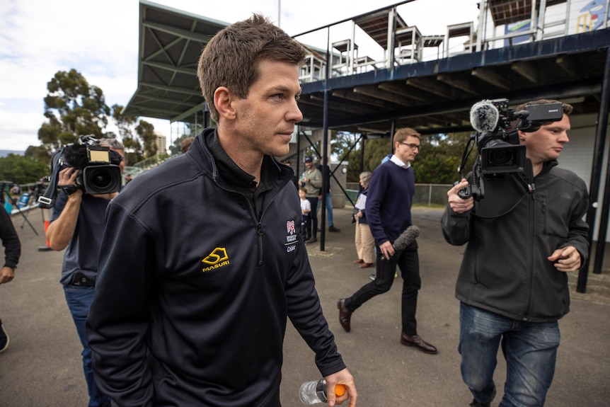 An Australian cricketer walks out of a cricket ground surrounded by TV cameramen after his game is called off.