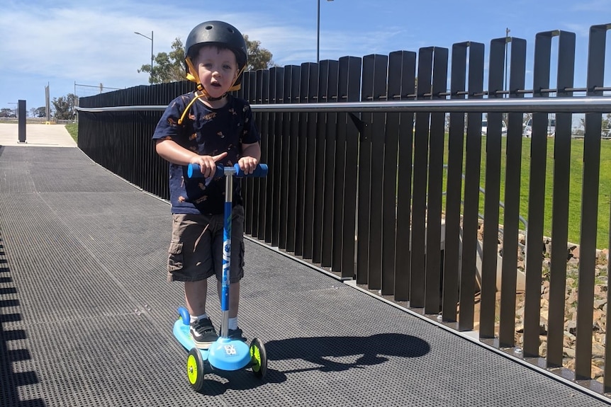 A little boy wearing a helmet rides a scooter along the footpath.