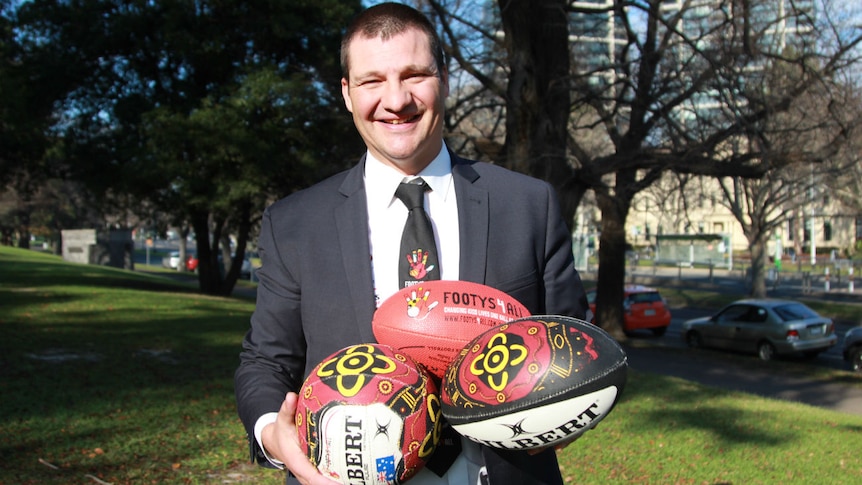 A man in a suit stands in a park holding an AFL football, netball and rugby ball painted with indigenous designs.