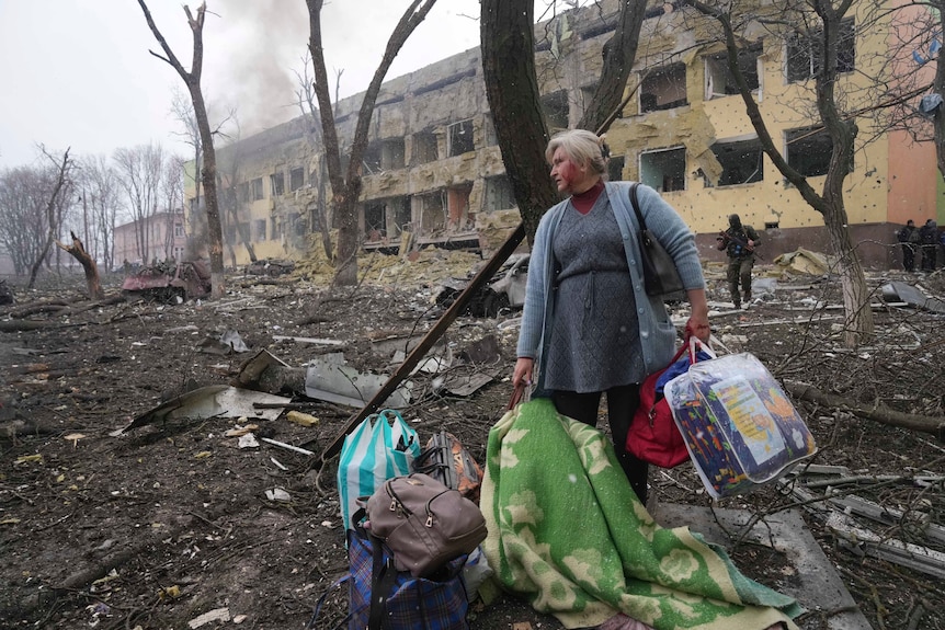 A woman stands near bags of luggage outside a large damaged building.