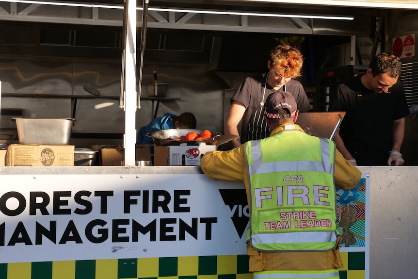 Firefighter gets food from food truck, forest fire management written on the front. Two people in black work behind the counter.