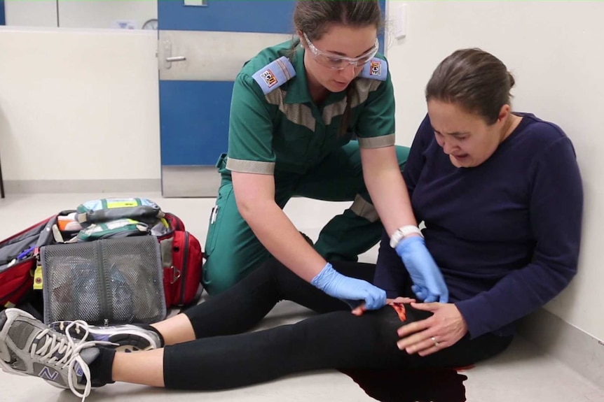 A student paramedic treats a woman seated on the floor with a bloody leg wound.