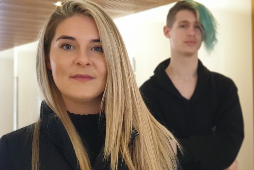 A young woman with long hair smiles with a man standing just behind her.