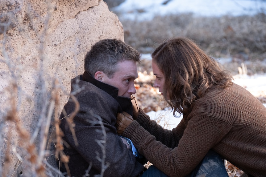 A white man is slouched against a rock in a desert, looking distressed. A white woman kneels before him, grabbing his lapel