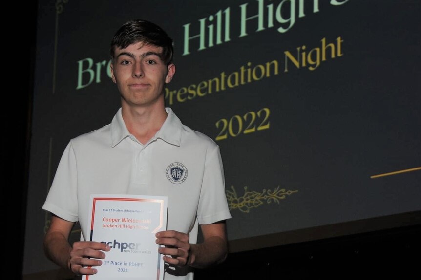 A teenager wearing a white school uniform holding a certificate standing in front of a projector screen