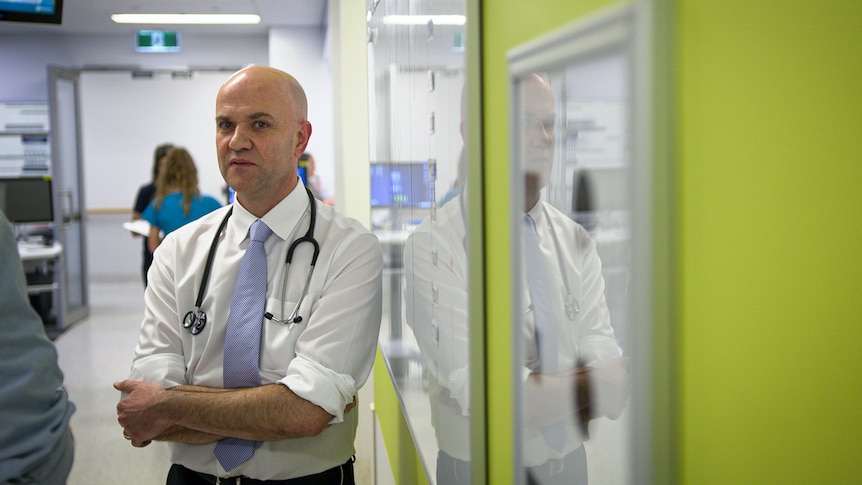 A doctor, looking stern with a stethoscope around his neck, stands in a hospital hallway.