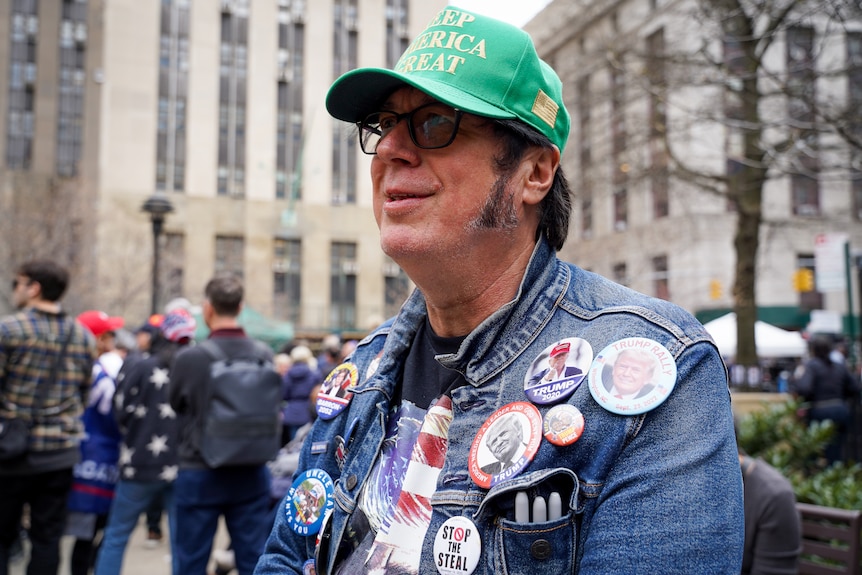 A man wearing a green truckers hat and a ton of Donald Trump badges on his denim jacket sits on a bench