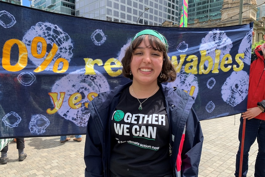 A smiling young girl with short brown hair stands in front of a sign about renewables