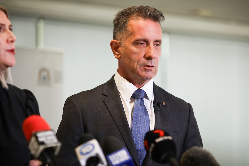 Paul Papalia wears a black suit jacket and navy tie, he stands behind a row of microphones.