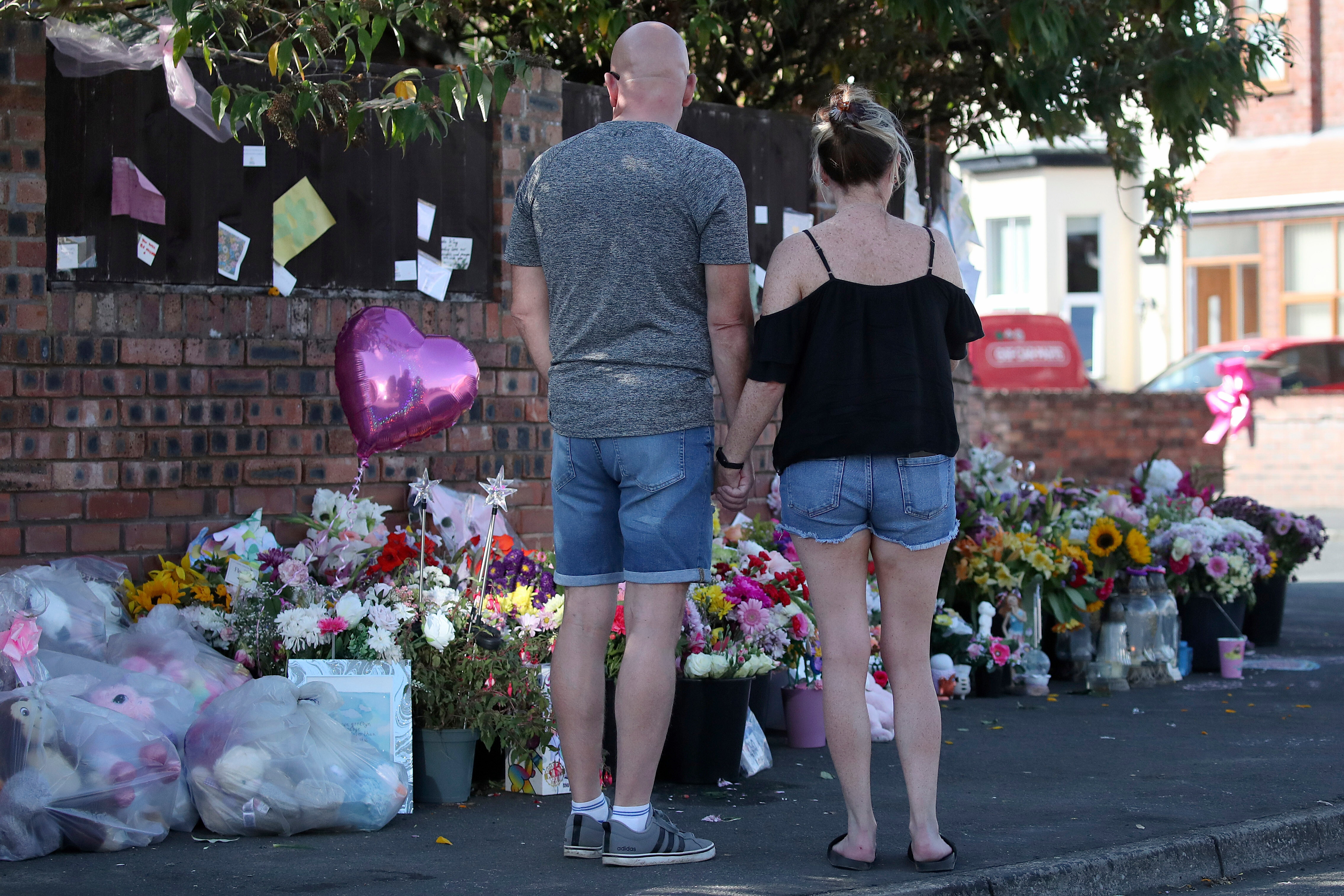 A man and a woman hold hands, as they stand looking at floral tributes and balloons at the side of a pavement.