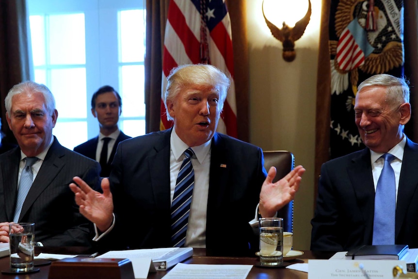 Donald Trump, flanked by Rex Tillerson and James Mattis, sits at a desk with flags on the wall in the background.