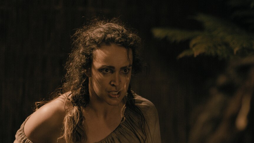 A woman with long dark curly hair and fierce expression in colonial blouse stands leaning forward ready for a confrontation.