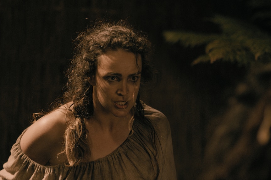 A woman with long dark curly hair and fierce expression in colonial blouse stands leaning forward ready for a confrontation.