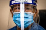 A woman wearing a blue face mask and a face shield looks seriously at the camera.
