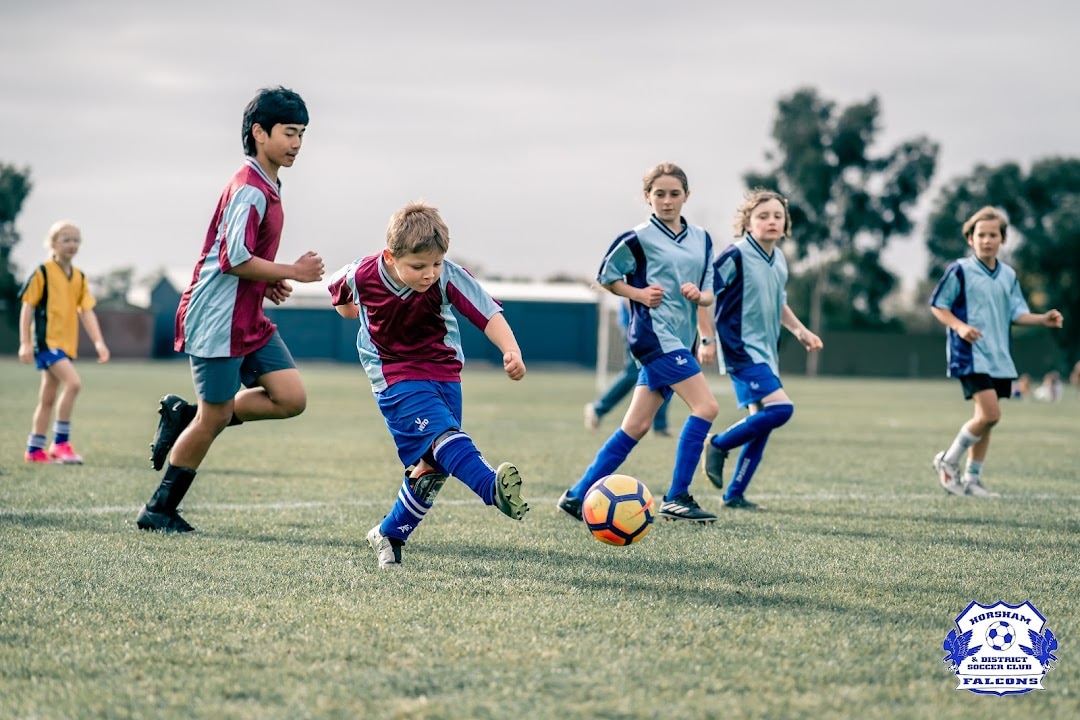 A young boy in a blue soccer jersey kicks a soccer ball while being pursued by other children 