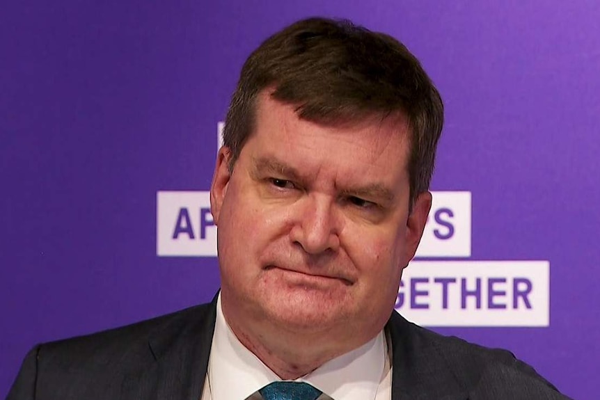 A man in a suit standing in front of a purple background.