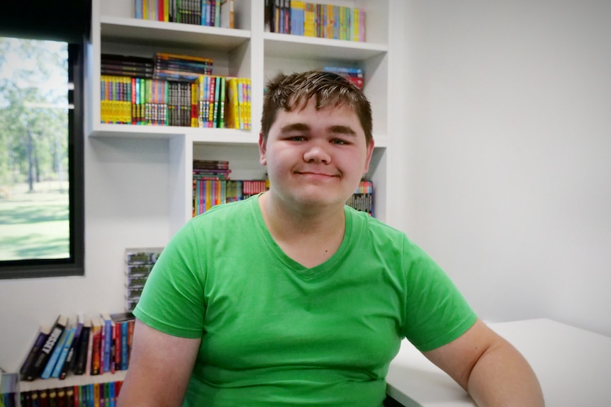 A teenage boy in a green shirt smiles at the camera, behind him is a bookshelf with colourful books.